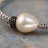 Macro close-up of the Killini bracelet — large baroque freshwater pearl with iridescent nacre, two stacked red sapphire pavé rondelles at its base, light grey macramé cord on both sides, on weathered blue-grey painted stone — JLL Pearl