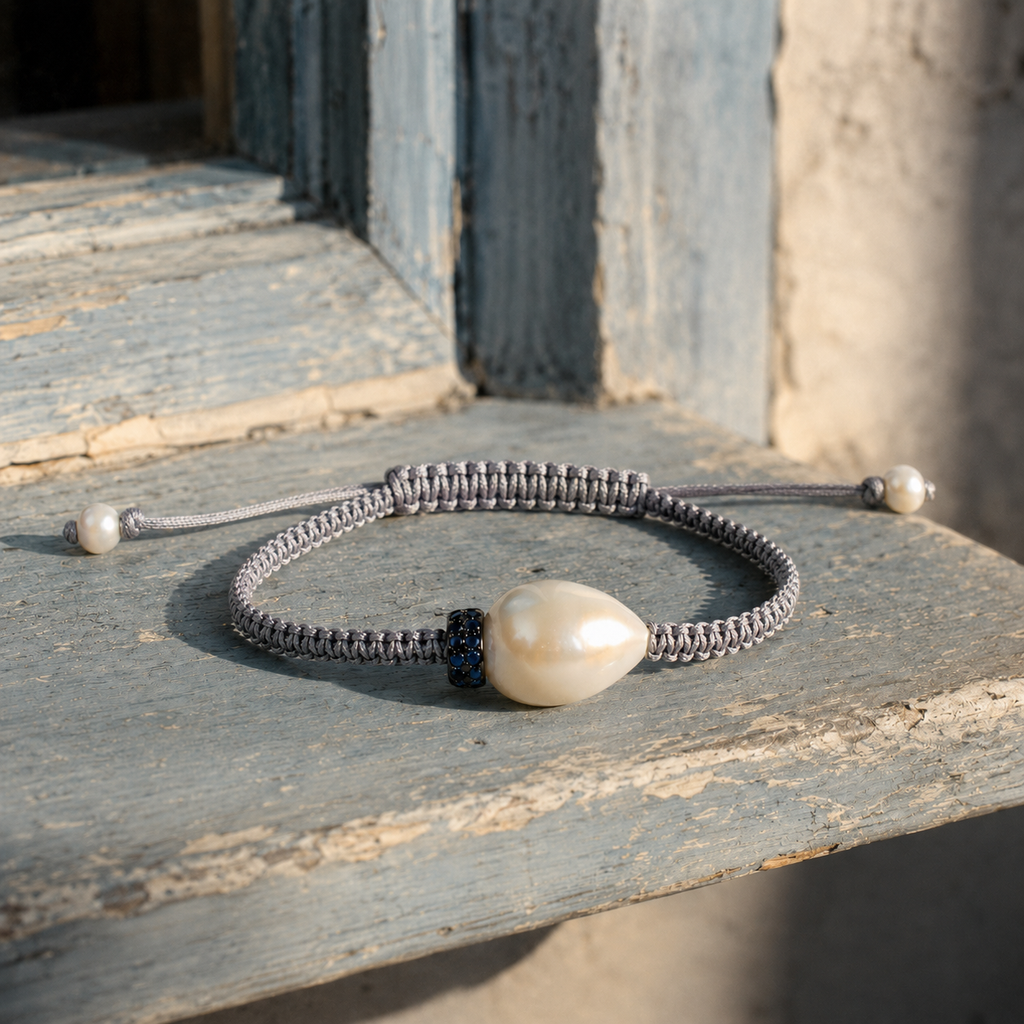 Killini bracelet — grey macramé cord, cream-white baroque pearl and blue sapphire rondelle — resting on aged blue-grey painted wood in Mediterranean light — JLL Pearl