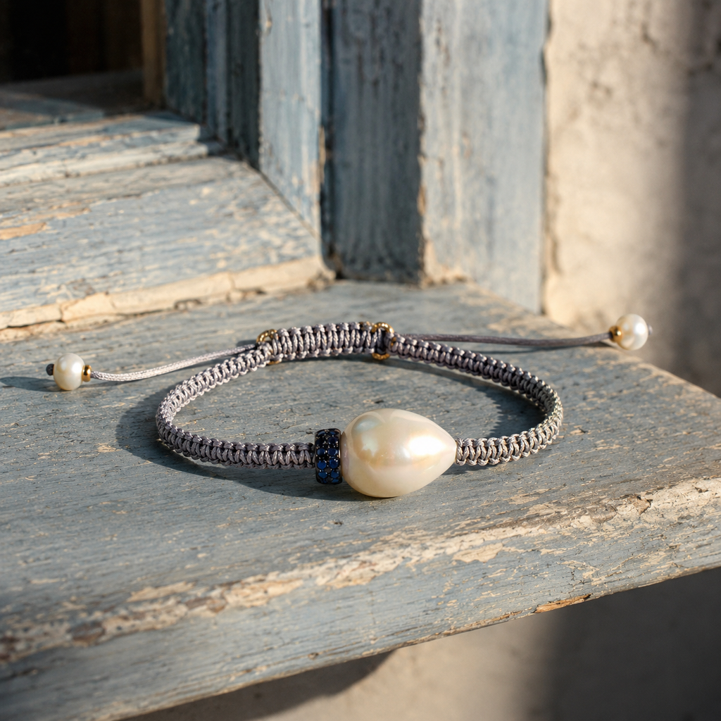 Killini bracelet — grey macramé cord, cream-white baroque pearl and blue sapphire rondelle — resting on aged blue-grey painted wood in Mediterranean light — JLL Pearl
