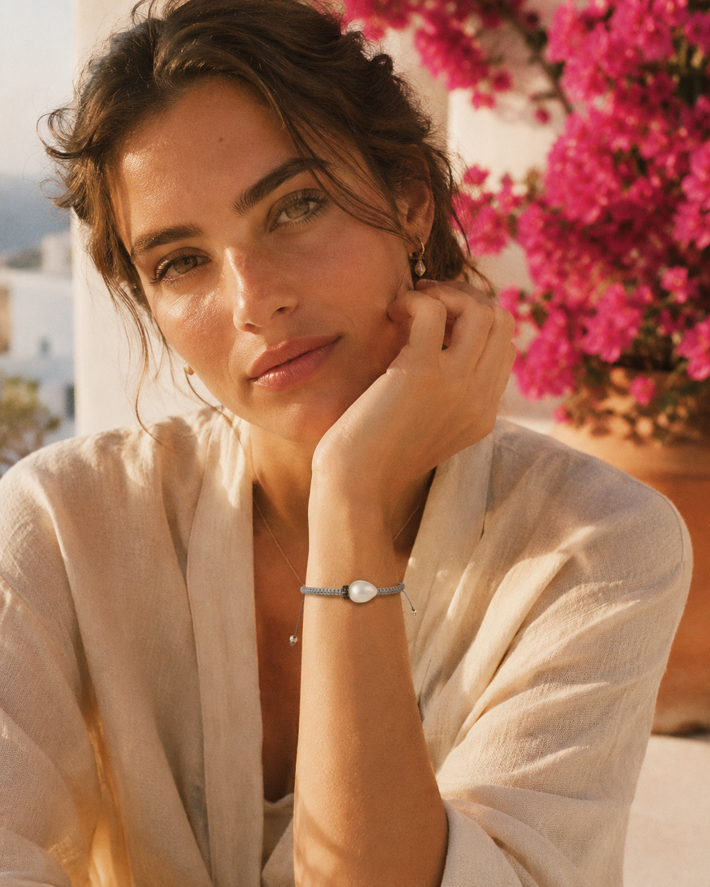 Young Mediterranean woman wearing the Killini grey macramé and baroque pearl bracelet, hand near face, pink bougainvillea in the background, golden hour — JLL Pearl