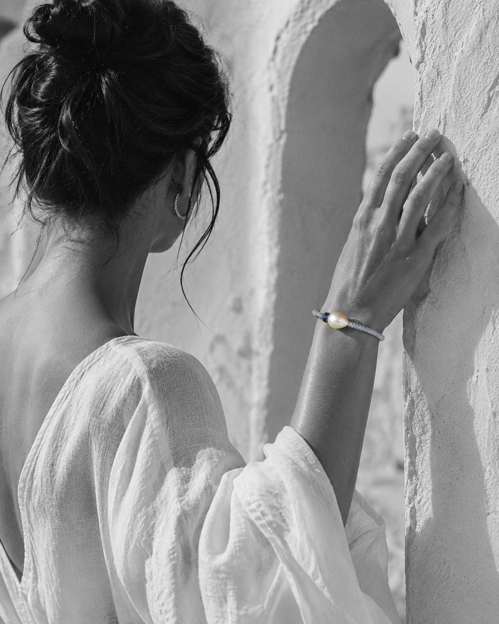 Killini grey macramé bracelet with baroque pearl and blue sapphire toupie on a Mediterranean woman's wrist, raised against a white Greek archway — JLL Pearl