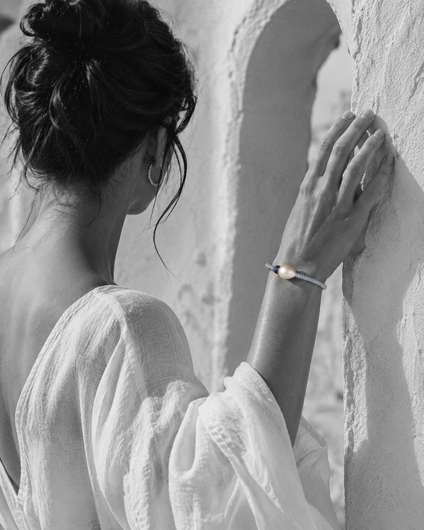 Killini grey macramé bracelet with baroque pearl and blue sapphire toupie on a Mediterranean woman's wrist, raised against a white Greek archway — JLL Pearl