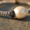 Macro close-up of the Killini bracelet: blue sapphire toupie in dark metal setting pressed against a drop-shaped cream-white baroque pearl, grey macramé cord — JLL Pearl