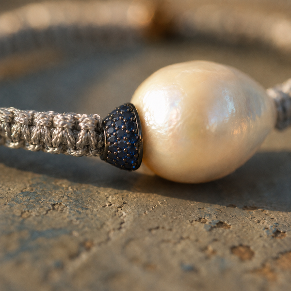 Macro close-up of the Killini bracelet: blue sapphire toupie in dark metal setting pressed against a drop-shaped cream-white baroque pearl, grey macramé cord — JLL Pearl