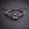 Kos amethyst topaz woven bracelet — flatlay on dark grey slate: pale lavender emerald-cut amethyst, pink topaz halo with gold beads, iridescent multicolour macramé cord — JLL Pearl