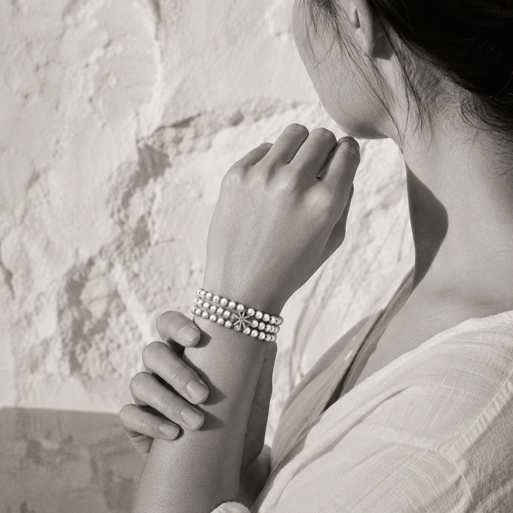 Lalibella bracelet worn on the wrist — three rows of round freshwater pearls knotted in grey satin cord, central 8-pointed diamond star, woman's hands cradling her wrist against a white Greek stone wall, black and white with selective colour on the bracelet — JLL Pearl