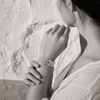 Lalibella bracelet worn on the wrist — three rows of round freshwater pearls knotted in grey satin cord, central 8-pointed diamond star, woman's hands cradling her wrist against a white Greek stone wall, black and white with selective colour on the bracelet — JLL Pearl