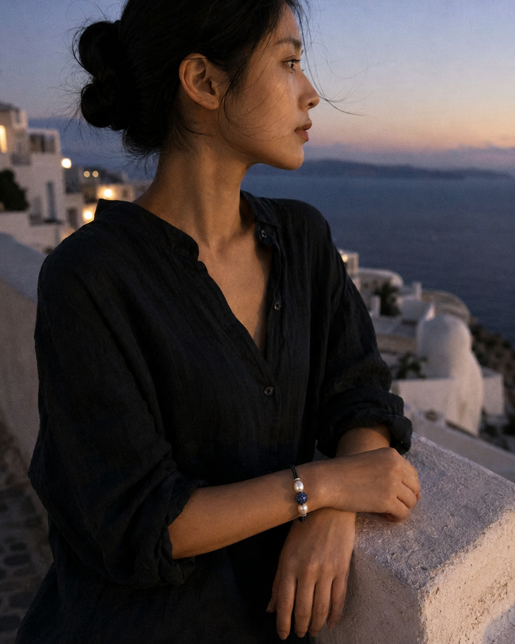 Manhattan bracelet by JLL Pearl — East Asian woman in ivory linen shirt, forearms on whitewashed Santorini railing at sunset, dark macramé bracelet with baroque pearls and blue sapphire ball clearly visible