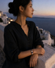 Manhattan bracelet by JLL Pearl — East Asian woman in ivory linen shirt, forearms on whitewashed Santorini railing at sunset, dark macramé bracelet with baroque pearls and blue sapphire ball clearly visible