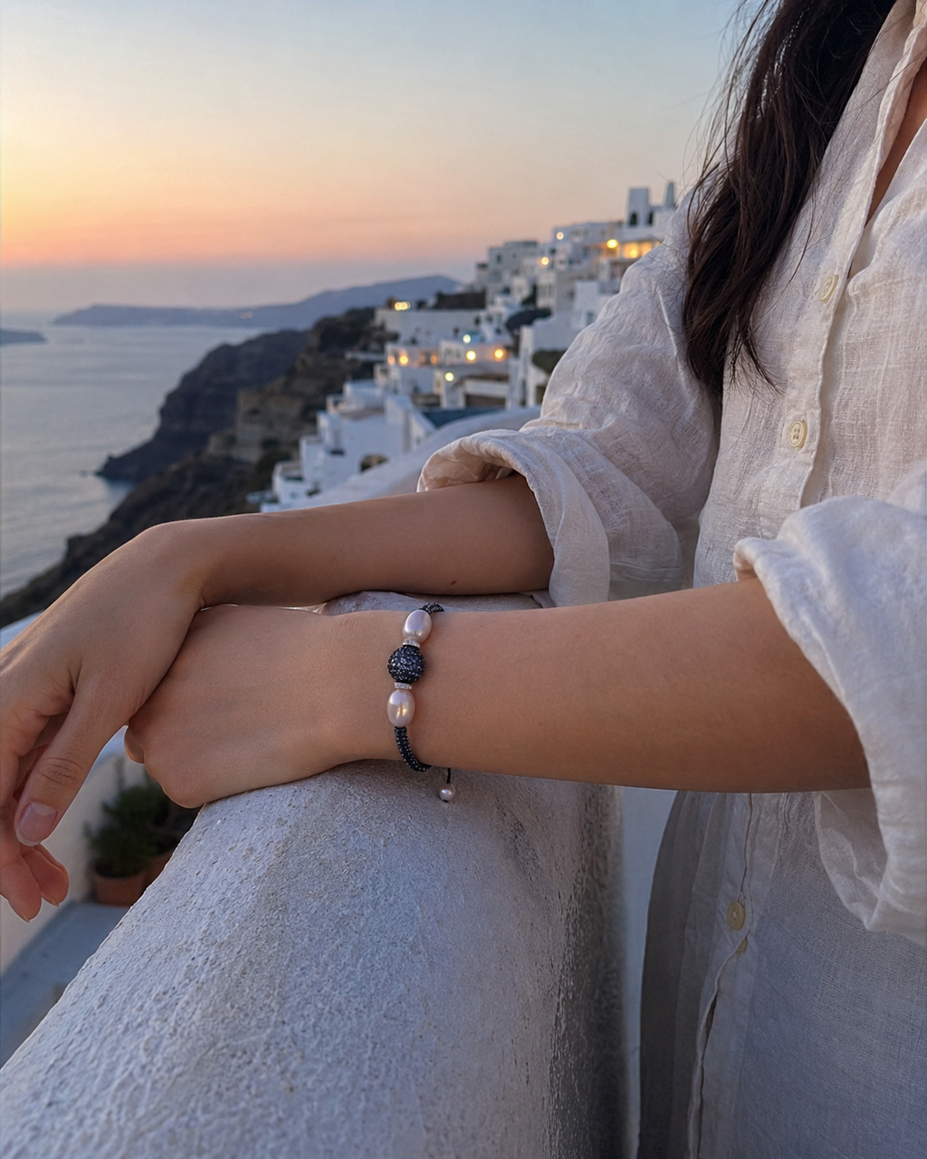 Manhattan bracelet by JLL Pearl — East Asian woman in ivory linen shirt, forearms on whitewashed Santorini railing at sunset, dark macramé bracelet with baroque pearls and blue sapphire ball clearly visible