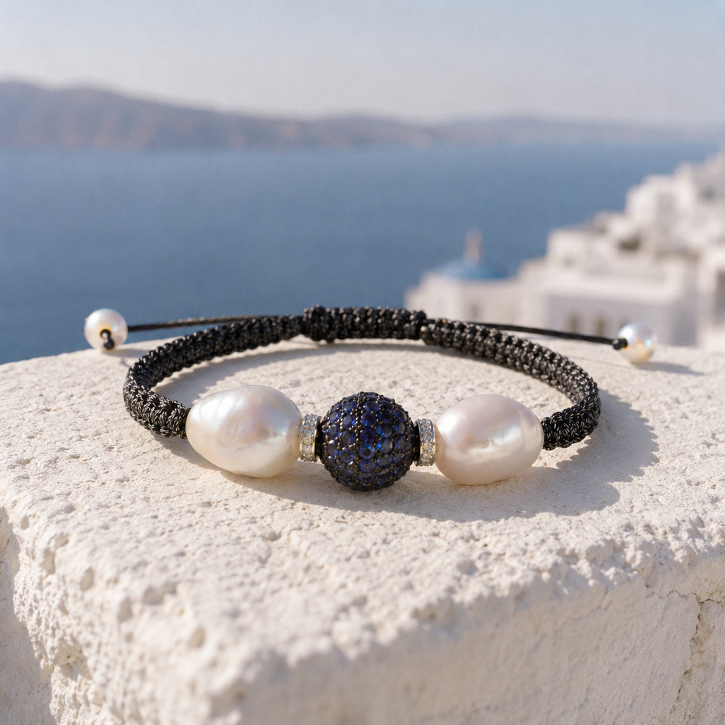 Manhattan bracelet by JLL Pearl — dark macramé cord with two baroque white freshwater pearls and pavé blue sapphire ball, resting on whitewashed Greek stone wall, Santorini caldera in background