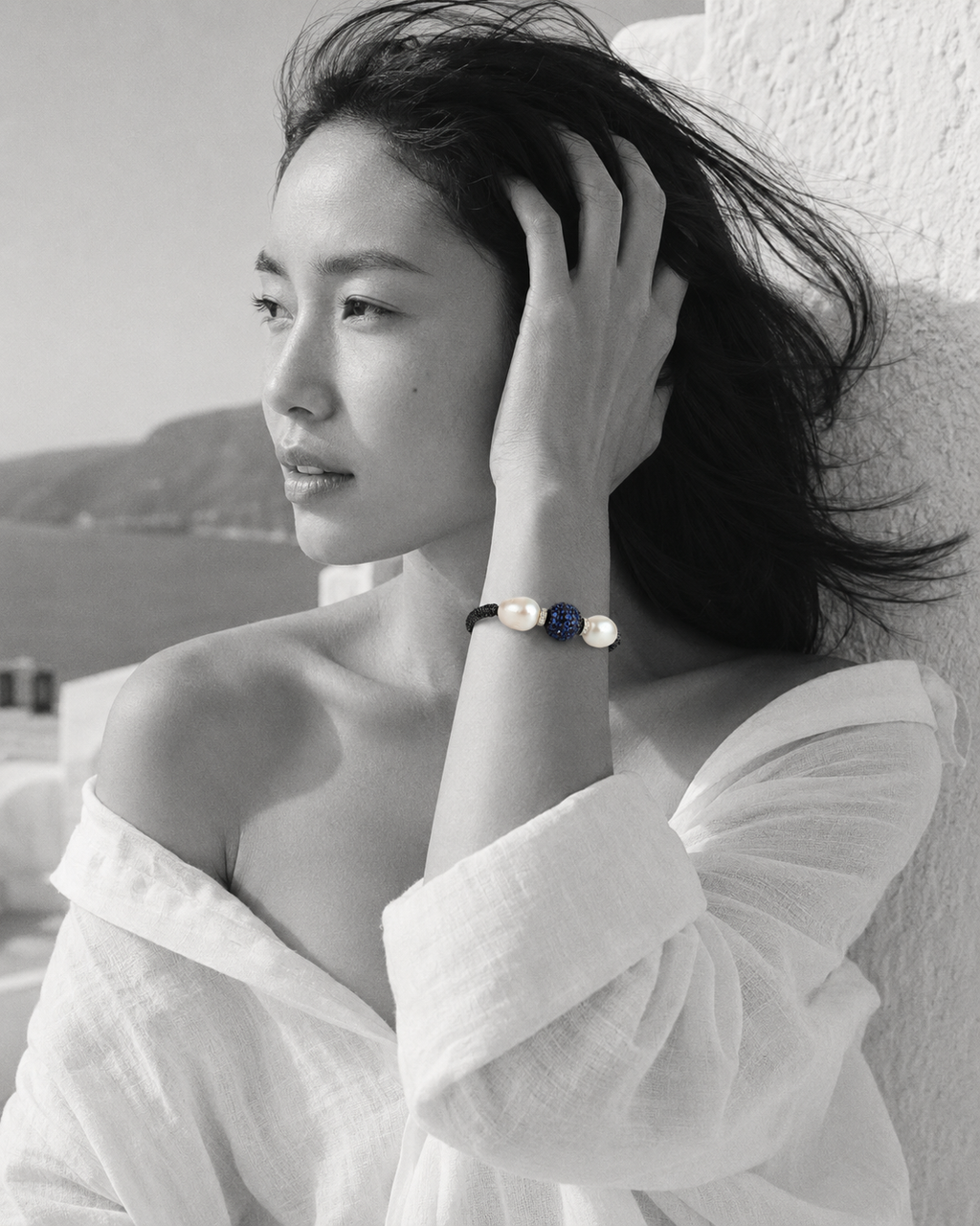 Manhattan bracelet by JLL Pearl — black and white portrait, East Asian woman, wind in hair, selective colour on dark macramé cord, baroque white pearls and deep blue sapphire ball
