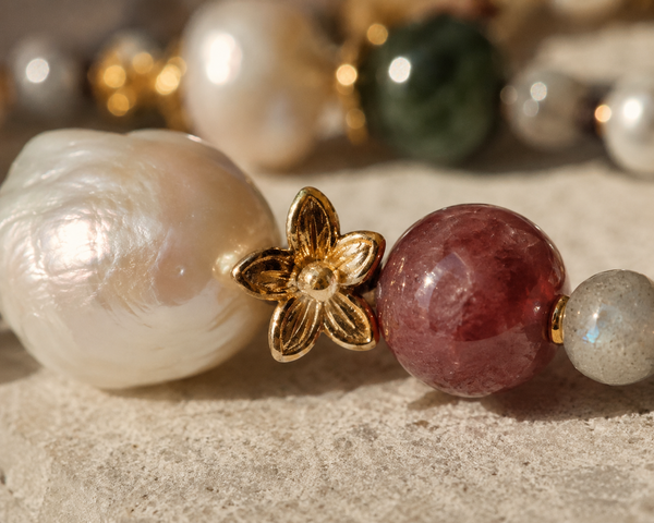 Miami baroque pearl necklace detail, gold flower spacer between large baroque freshwater pearl and red-brown tourmaline, labradorite visible at right, macro on pale sandstone, handmade by JLL Pearl