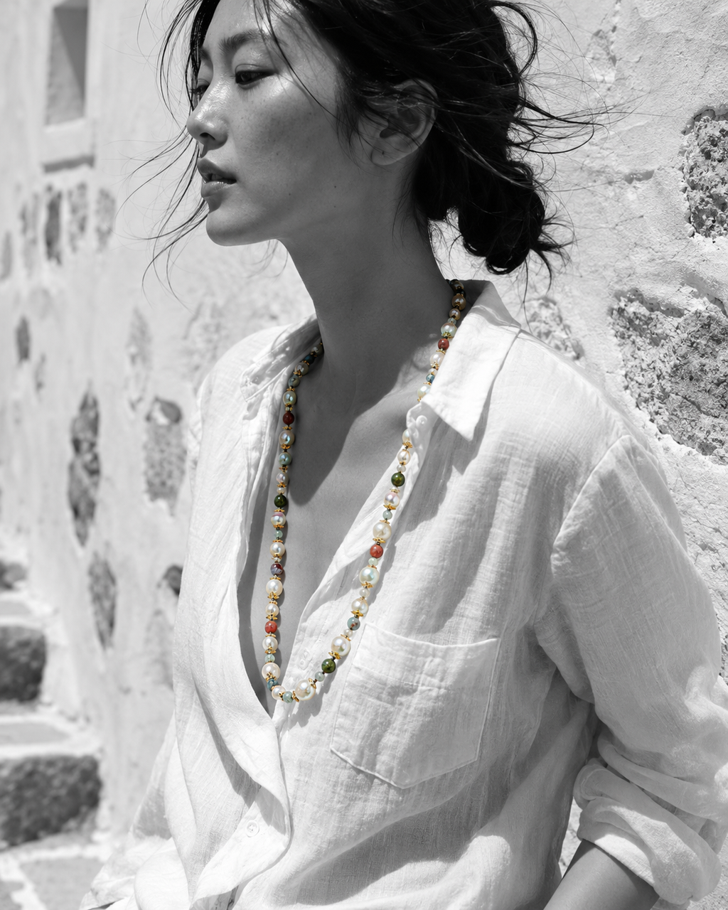 Miami baroque pearl necklace with tourmaline and labradorite worn by Asian model in profile, white linen shirt, whitewashed stone wall, wind in hair, natural colour necklace on black and white image, handmade by JLL Pearl