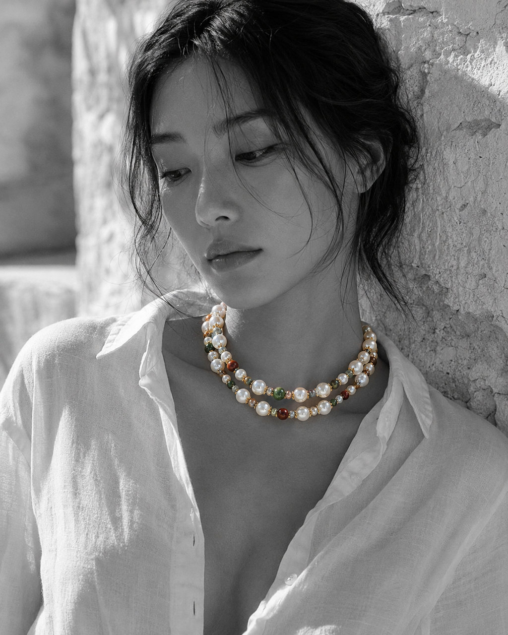 Miami baroque pearl necklace with tourmaline worn doubled as two-strand choker at throat by Asian model, whitewashed Greek stone wall, white linen shirt, natural colour necklace on black and white image, handmade by JLL Pearl