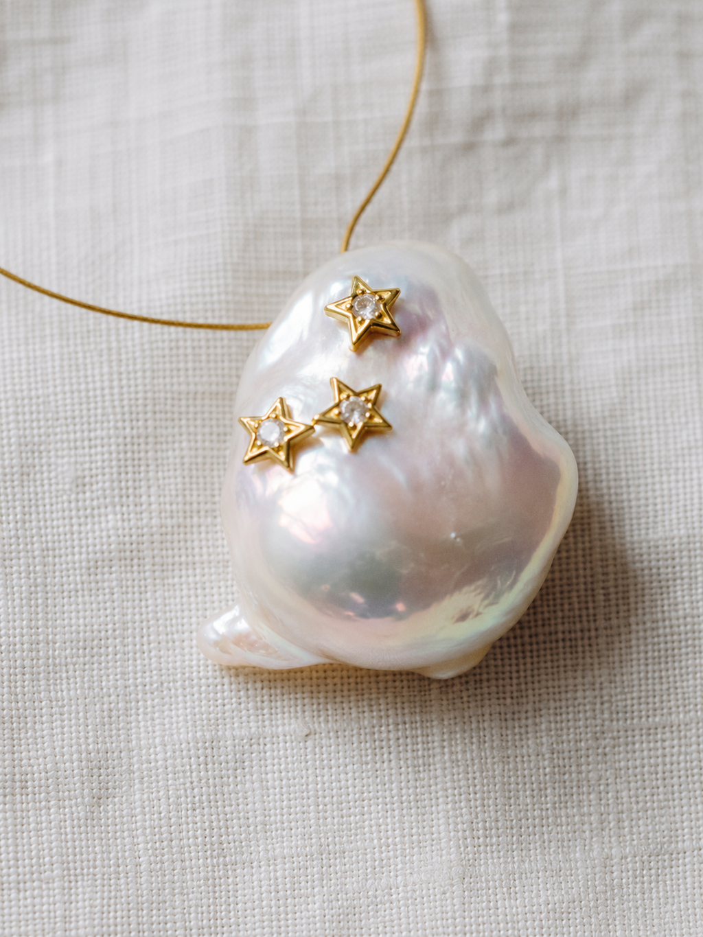 Close-up of Mila baroque pearl pendant with three gold star ornaments set with white topaz, iridescent freshwater baroque pearl on pale linen, JLL Pearl