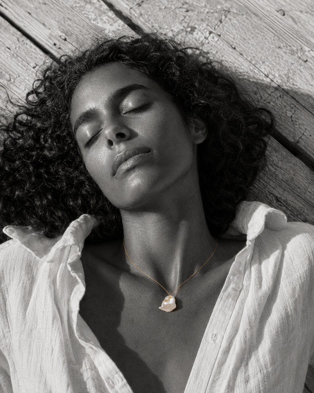 Mila baroque pearl white topaz gold necklace worn by a woman lying on sun-bleached wooden pier planks, black and white editorial photograph with selective color on jewelry, JLL Pearl