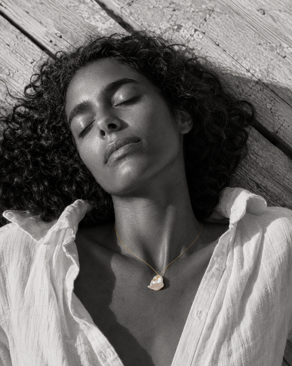 Mila baroque pearl white topaz gold necklace worn by a woman lying on sun-bleached wooden pier planks, black and white editorial photograph with selective color on jewelry, JLL Pearl