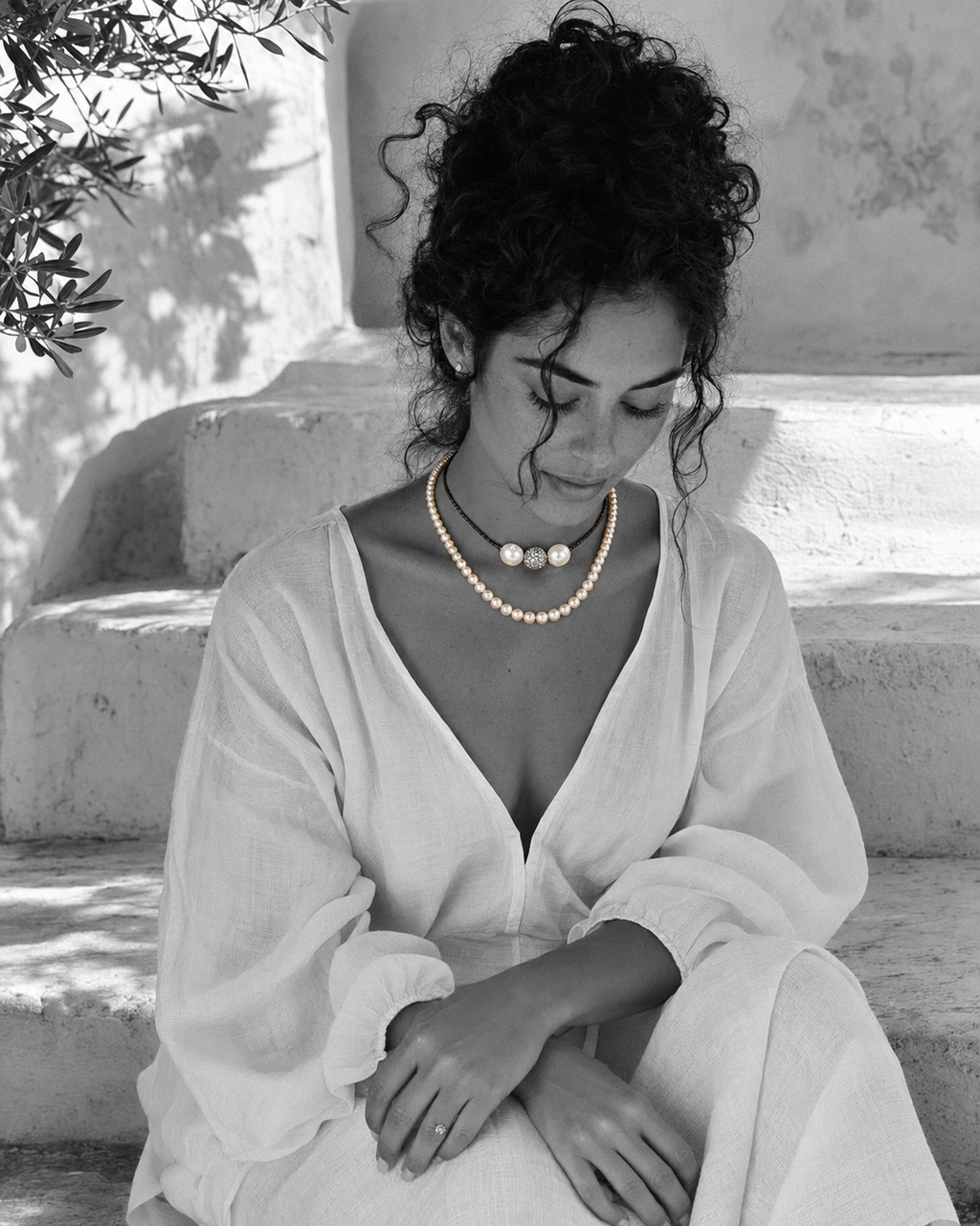 Mina pearl spinel white topaz necklace worn by a woman seated on Greek stone steps, black and white portrait with olive tree and whitewashed wall
