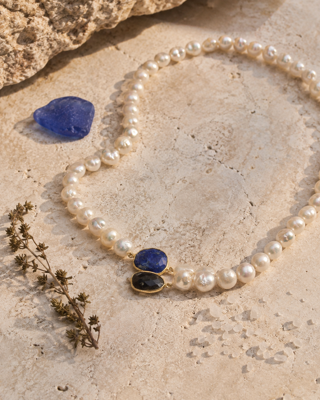 Nati pearl necklace lapis lazuli sapphire gold arranged on pale travertine stone with cobalt blue sea glass and dried thyme, JLL Pearl