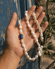 Nati pearl necklace lapis lazuli sapphire gold held in an open feminine palm, Mediterranean background with blue shutters and dark olives, JLL Pearl