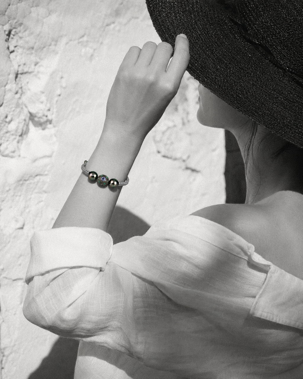 Black and white portrait of woman adjusting hat, Paros Tahitian pearl macramé bracelet in selective colour on wrist, Greek wall