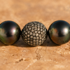 Macro close-up of the Paros bracelet — champagne diamond pavé ball flanked by two large dark Tahitian pearls with peacock iridescence, on warm terracotta stone — JLL Pearl