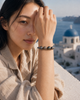 Paros Tahitian pearl and champagne diamond bracelet worn on the wrist of a young woman, hand raised near her face, Santorini blue-domed church and Aegean sea in the background — JLL Pearl