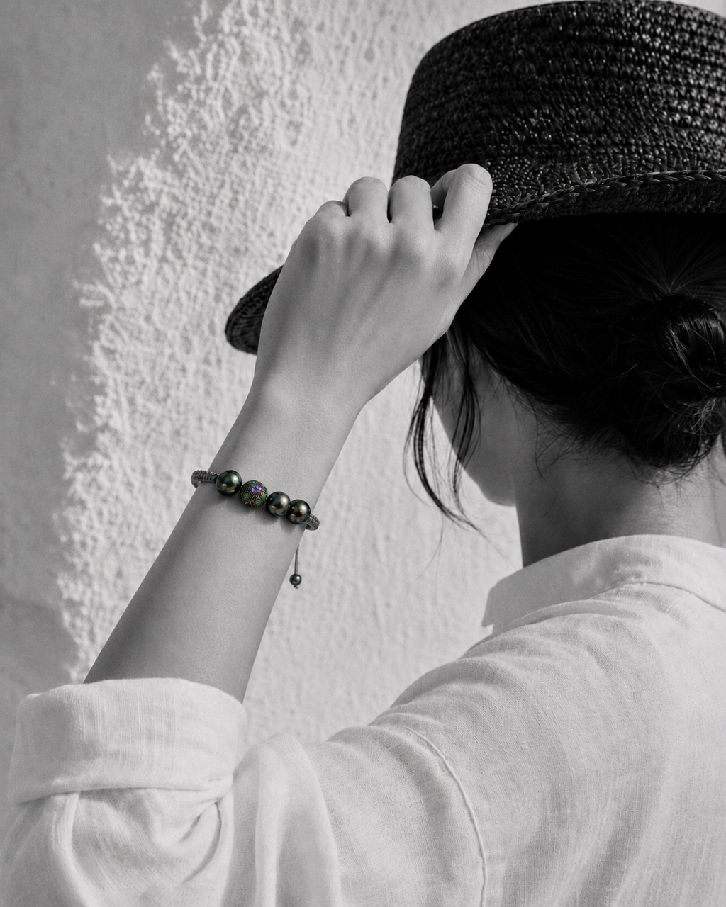 Paros Tahitian pearl macramé bracelet — four dark green-black Tahitian pearls and ornate tanzanite and emerald centre ball — worn on the wrist of a woman adjusting her hat against a white Greek wall, black and white with selective colour on the bracelet — JLL Pearl