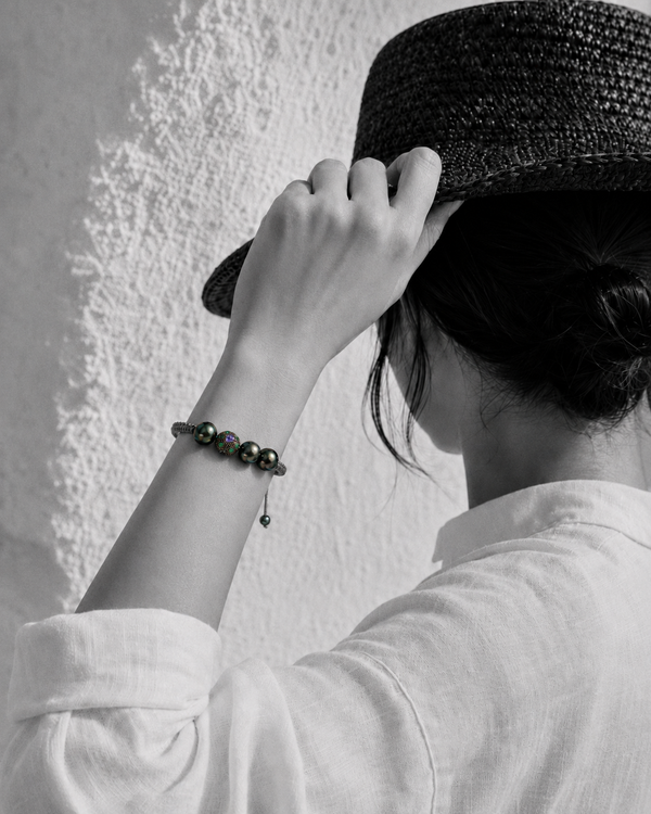 Paros Tahitian pearl macramé bracelet — four dark green-black Tahitian pearls and ornate tanzanite and emerald centre ball — worn on the wrist of a woman adjusting her hat against a white Greek wall, black and white with selective colour on the bracelet — JLL Pearl
