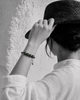 Paros Tahitian pearl macramé bracelet — four dark green-black Tahitian pearls and ornate tanzanite and emerald centre ball — worn on the wrist of a woman adjusting her hat against a white Greek wall, black and white with selective colour on the bracelet — JLL Pearl