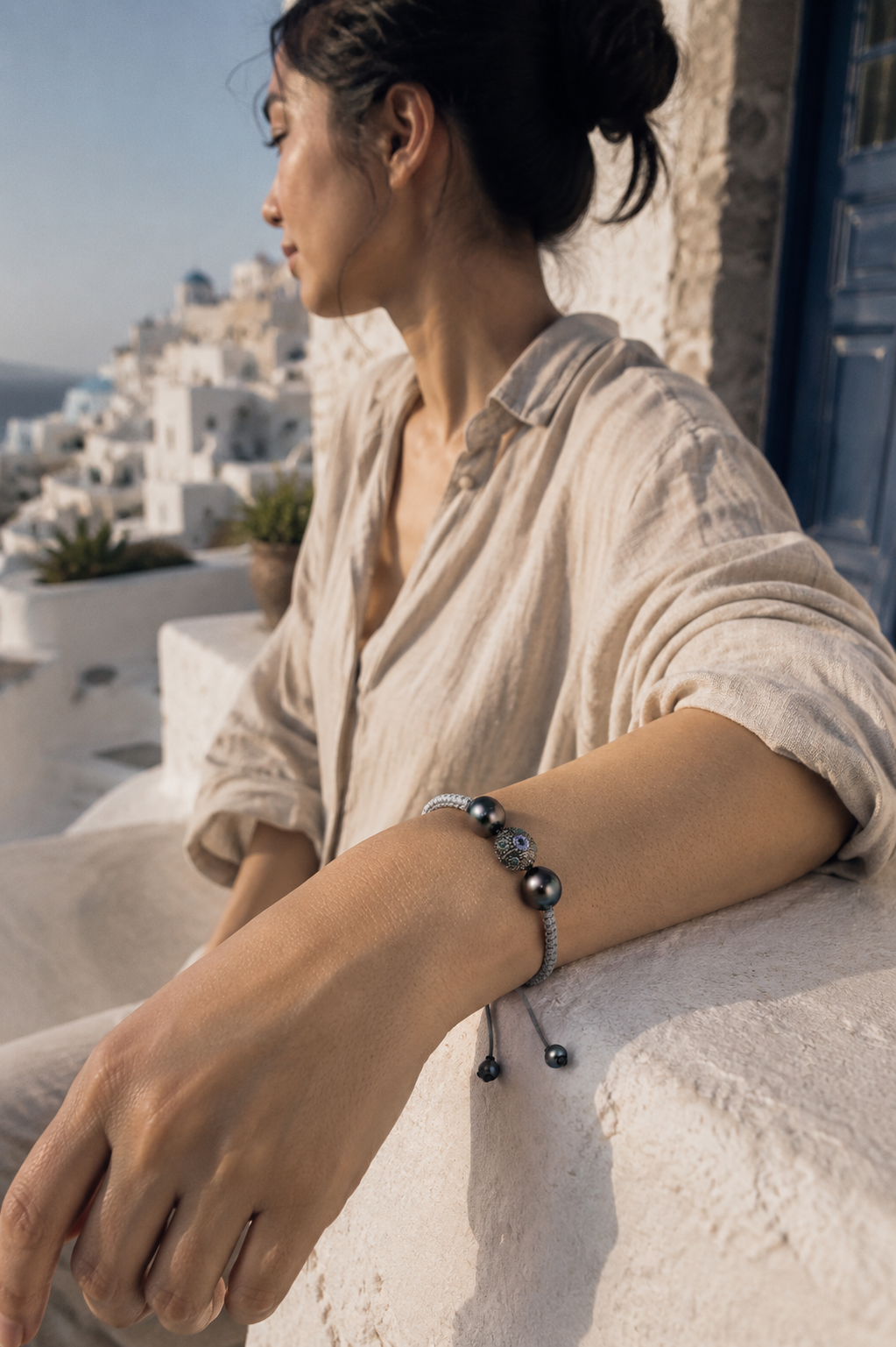 Woman wearing Paros Tahitian pearl macramé bracelet on terrace overlooking Santorini, natural light, linen shirt
