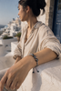 Woman wearing Paros Tahitian pearl macramé bracelet on terrace overlooking Santorini, natural light, linen shirt