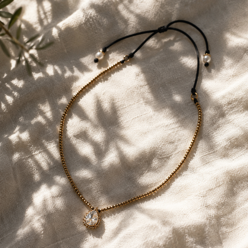 Romy white topaz pendant hematite necklace laid on white linen, gold hematite beaded chain with pear-cut topaz pendant, black satin cord with pearl and onyx finials, olive leaf shadows, handmade by JLL Pearl