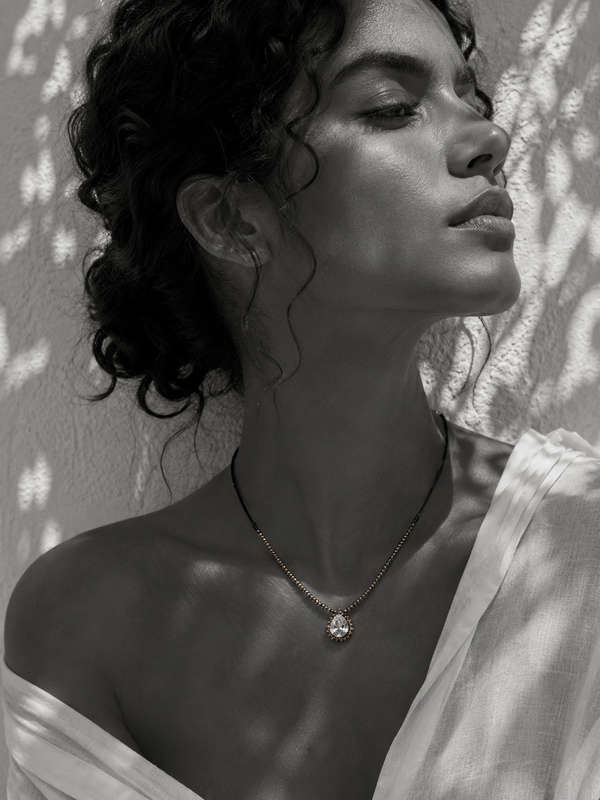 Romy white topaz pendant hematite necklace worn at the collarbone, pear-cut topaz in gold halo setting on gold hematite beaded chain, black satin cord, olive leaf shadows on whitewashed wall, handmade by JLL Pearl