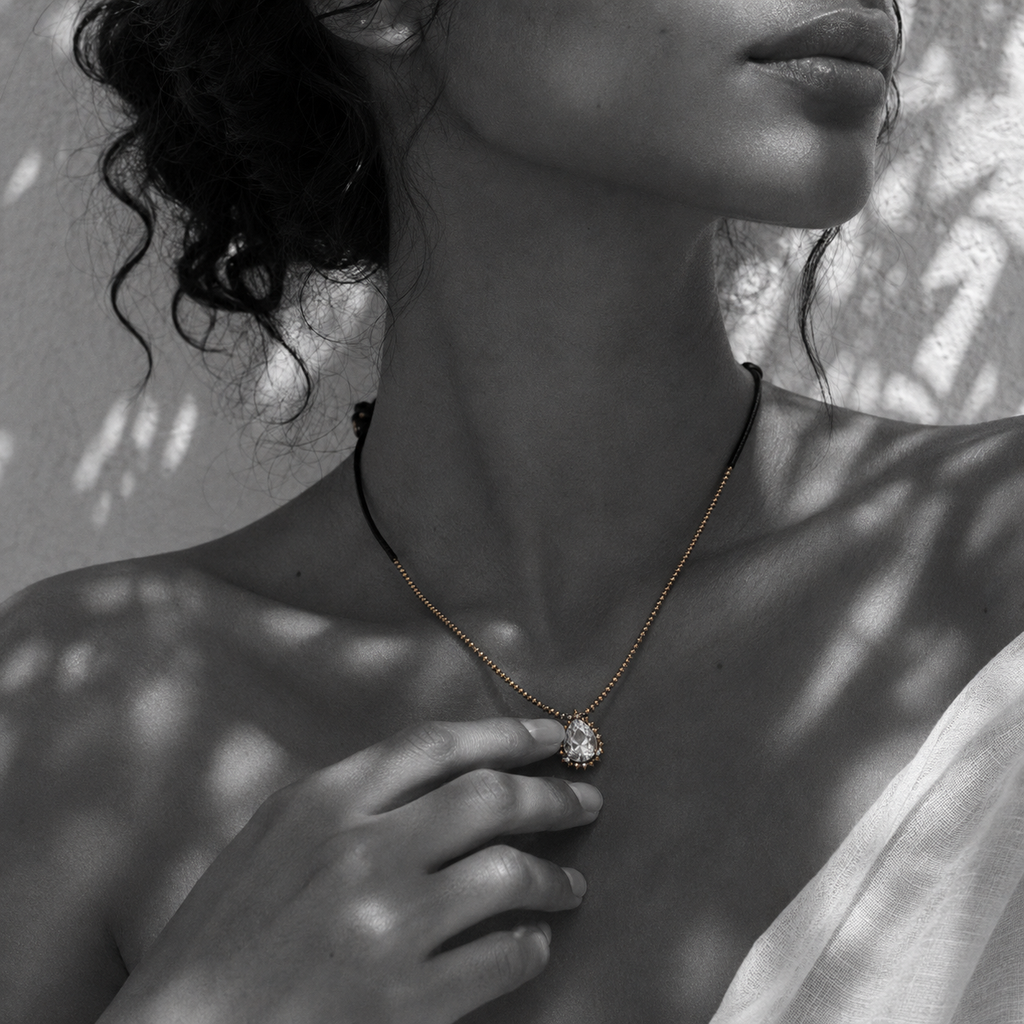 Hand touching Romy white topaz pendant hematite necklace at the collarbone, pear-cut topaz in gold halo, gold hematite beaded chain, black satin cord, olive leaf shadows on whitewashed wall, handmade by JLL Pearl