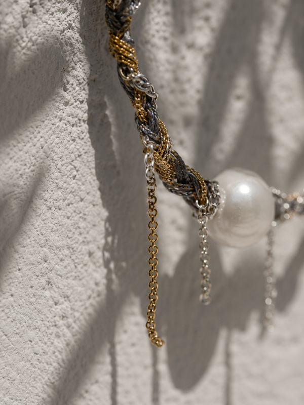 Close-up of Samos braided choker showing charcoal cord with gold and silver chains, free-hanging chain fringe and round freshwater pearl, olive leaf shadows on whitewashed plaster wall, handmade by JLL Pearl