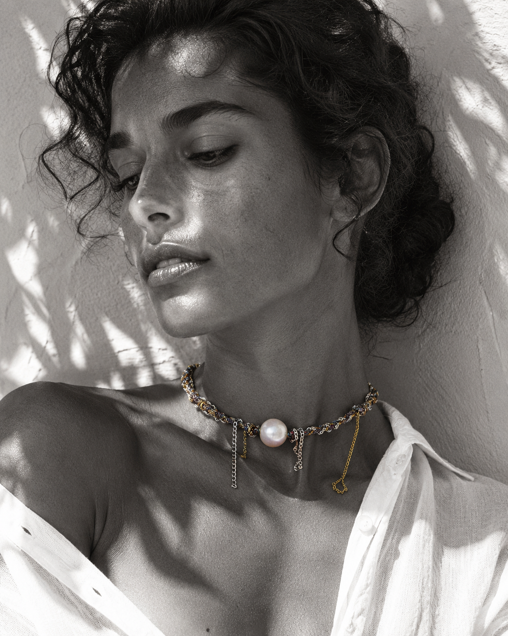 Samos braided silver gold chain pearl choker worn at the throat, round freshwater pearl at centre with hanging chain fringe, dark curly hair, white linen, olive leaf shadows on whitewashed wall, handmade by JLL Pearl