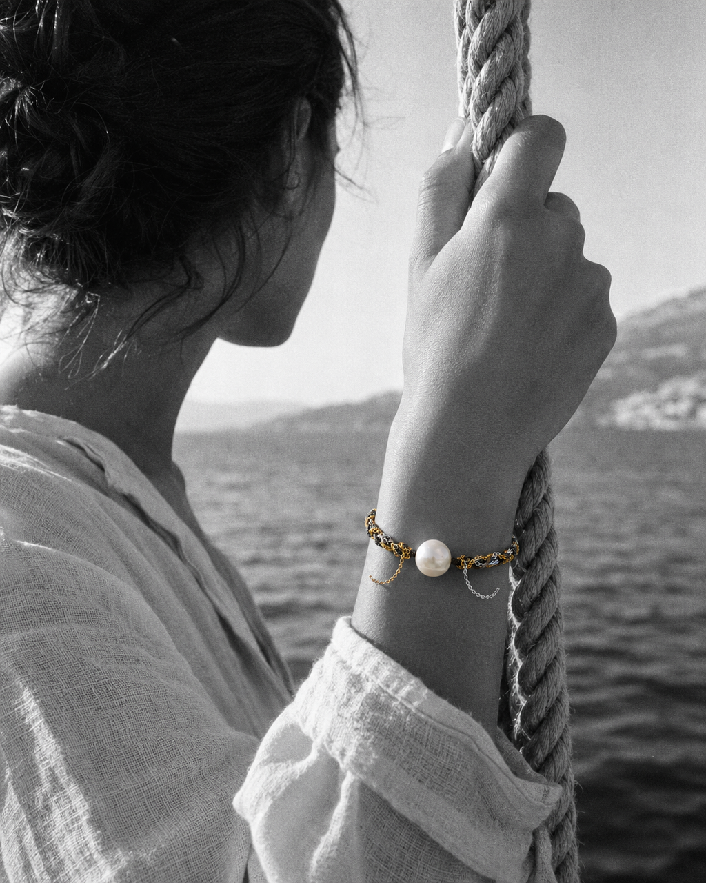 Samos freshwater pearl gold chain bracelet on a Mediterranean woman's wrist, hand gripping a braided sailing rope on a Greek caïque, Aegean sea in the background — JLL Pearl