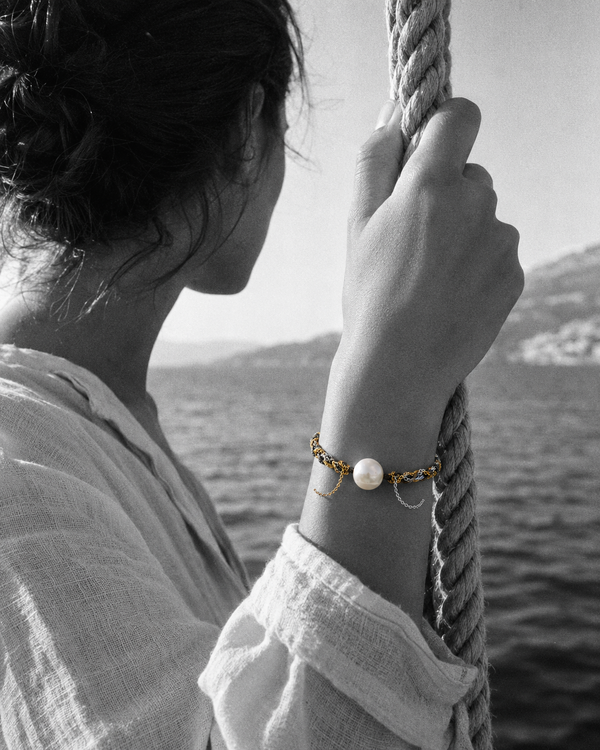 Samos freshwater pearl gold chain bracelet on a Mediterranean woman's wrist, hand gripping a braided sailing rope on a Greek caïque, Aegean sea in the background — JLL Pearl