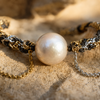 Macro close-up of the Samos bracelet: large round cream-white freshwater pearl at the centre of a braided gold, silver, and dark metal chain band, two fine hanging chains (one gold, one silver) — JLL Pearl