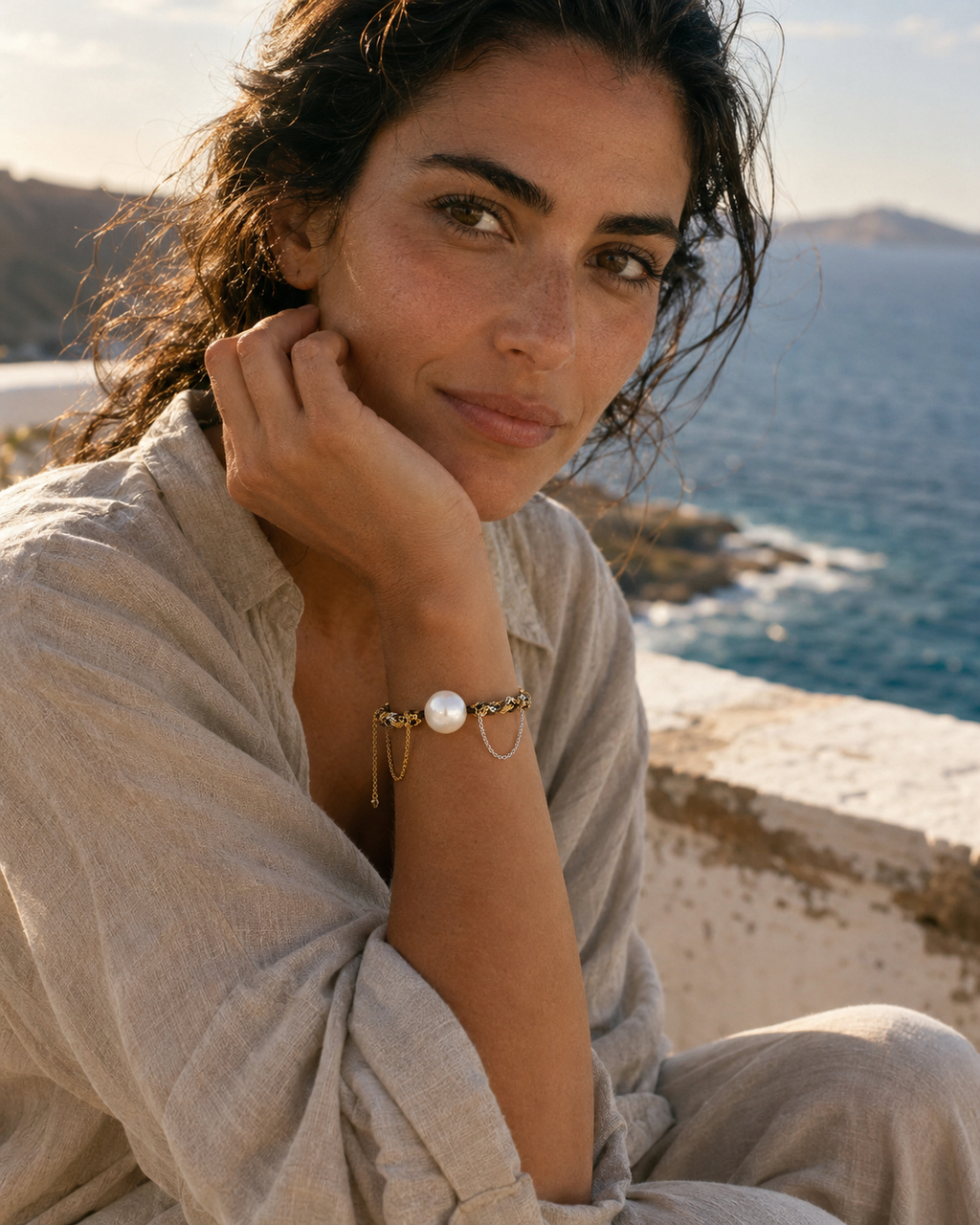Mediterranean woman, 30s, wearing the Samos freshwater pearl gold chain bracelet, hand near her face, seated on a whitewashed stone wall with the Aegean sea and Greek cliffs behind her — JLL Pearl