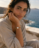 Mediterranean woman, 30s, wearing the Samos freshwater pearl gold chain bracelet, hand near her face, seated on a whitewashed stone wall with the Aegean sea and Greek cliffs behind her — JLL Pearl