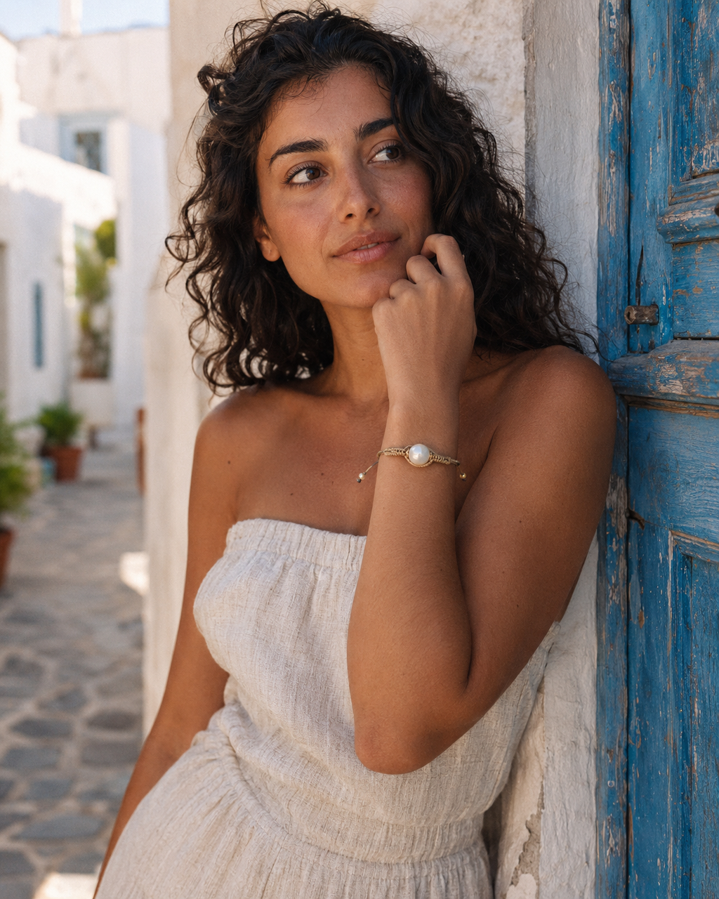 Tama bracelet worn by a Mediterranean woman leaning against a blue Greek door, hand near face, round pearl and gold macramé clearly visible
