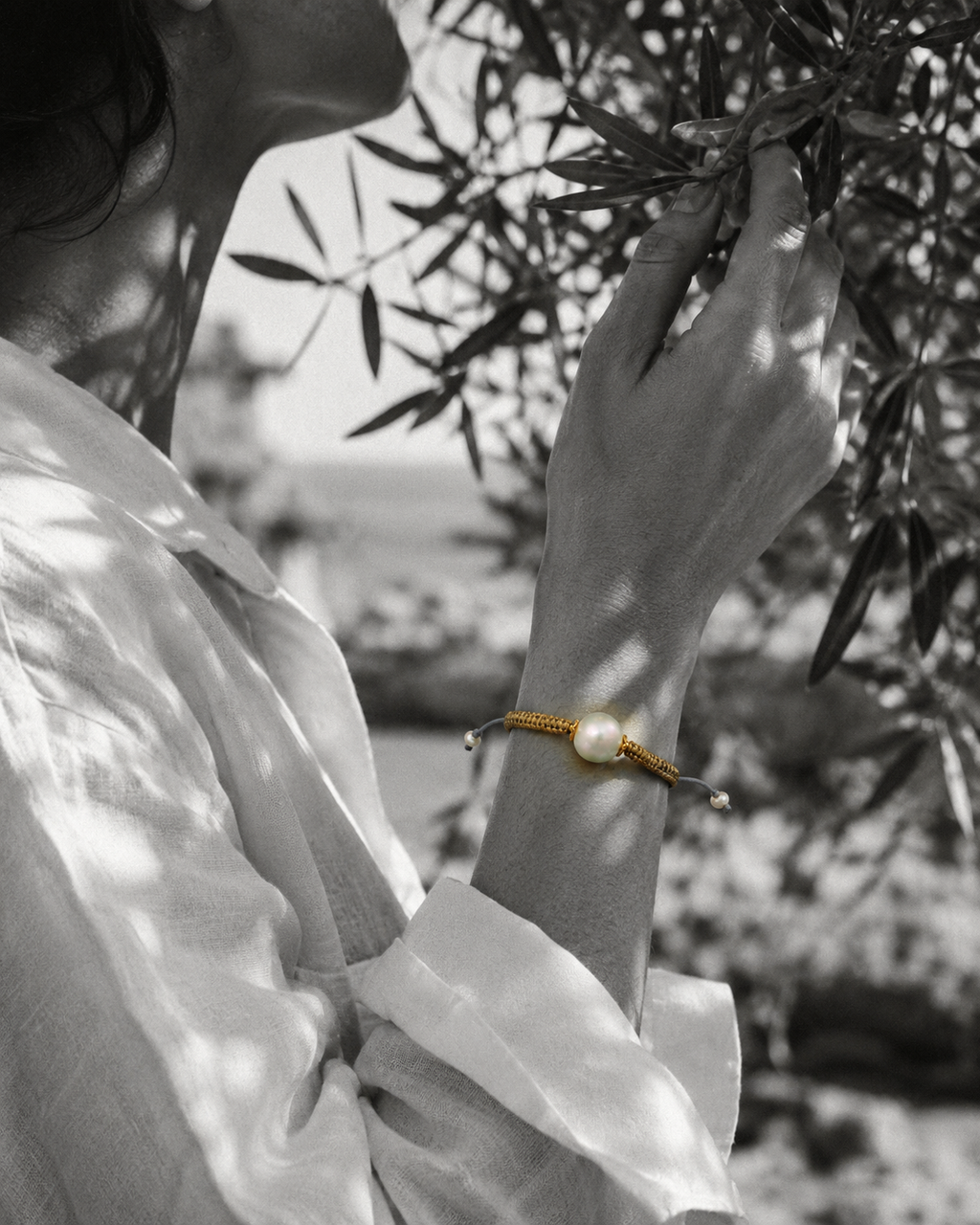 Tama bracelet — round freshwater pearl and gold macramé on wrist, olive branch background, black and white photograph with selective colour on bracelet