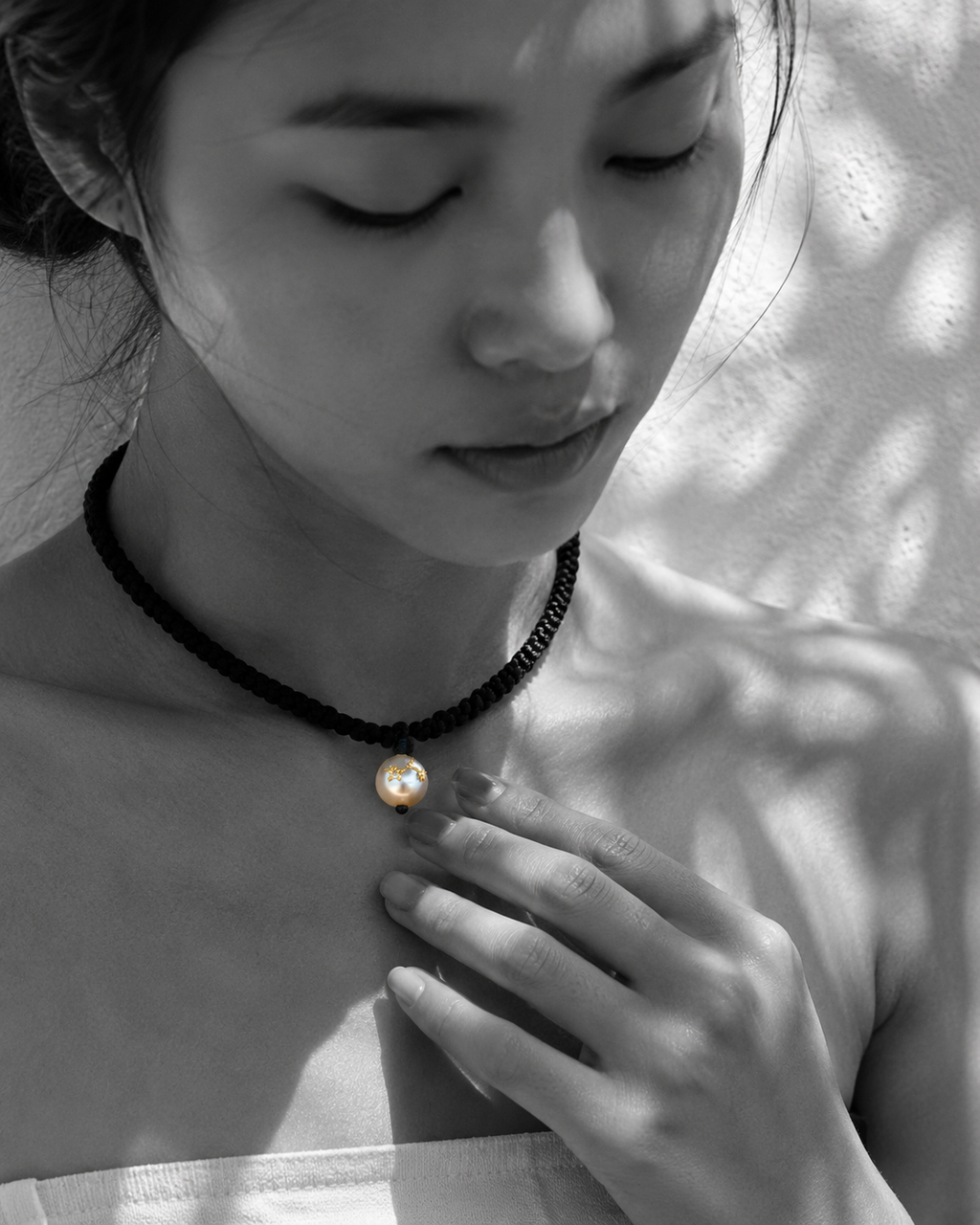 Tara freshwater pearl macramé choker necklace worn by an Asian model, hand resting on pearl with gold stars, dappled leaf shadows on whitewashed wall, B&W with pearl and gold stars in natural colour, handmade by JLL Pearl