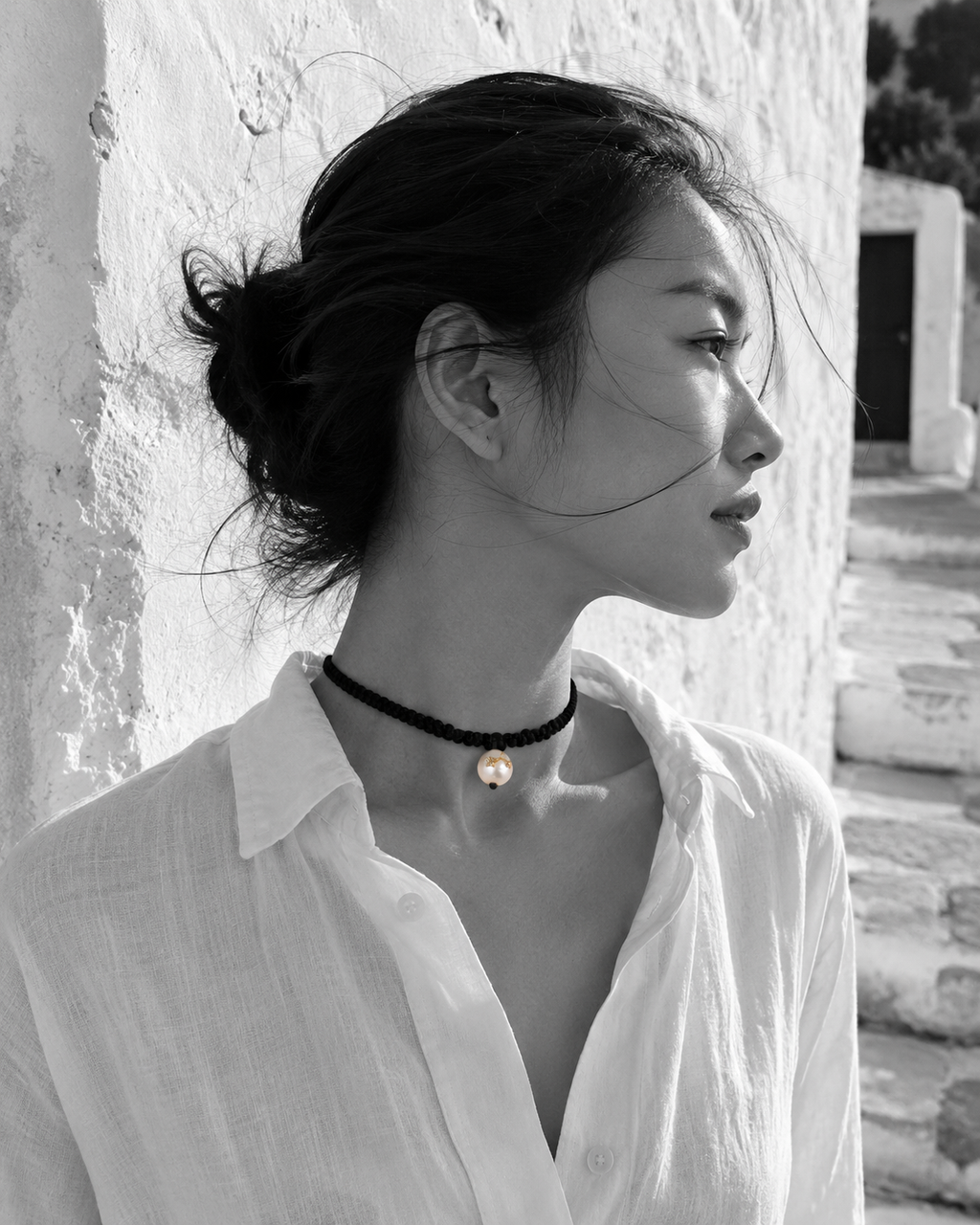 Tara freshwater pearl macramé choker necklace worn in profile by an Asian model against a whitewashed Greek wall, black satin braided cord, pearl with gold stars in natural colour, handmade by JLL Pearl