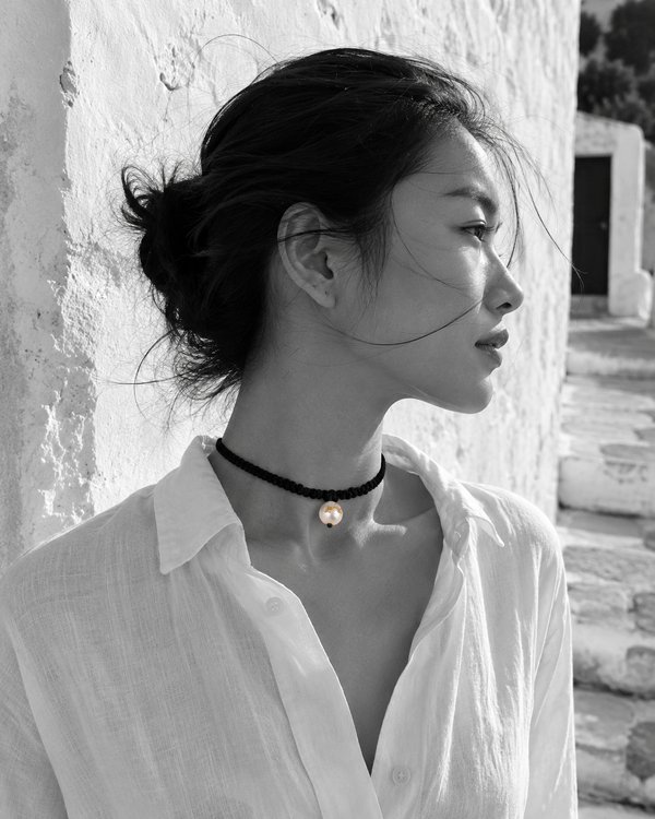 Tara freshwater pearl macramé choker necklace worn in profile by an Asian model against a whitewashed Greek wall, black satin braided cord, pearl with gold stars in natural colour, handmade by JLL Pearl