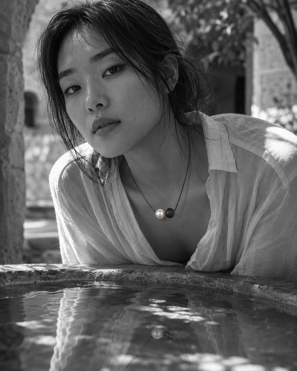 Zephyr pearl necklace black spinel oxidized silver lifestyle, Asian model leaning over stone fountain in Greek courtyard, necklace reflected in still water