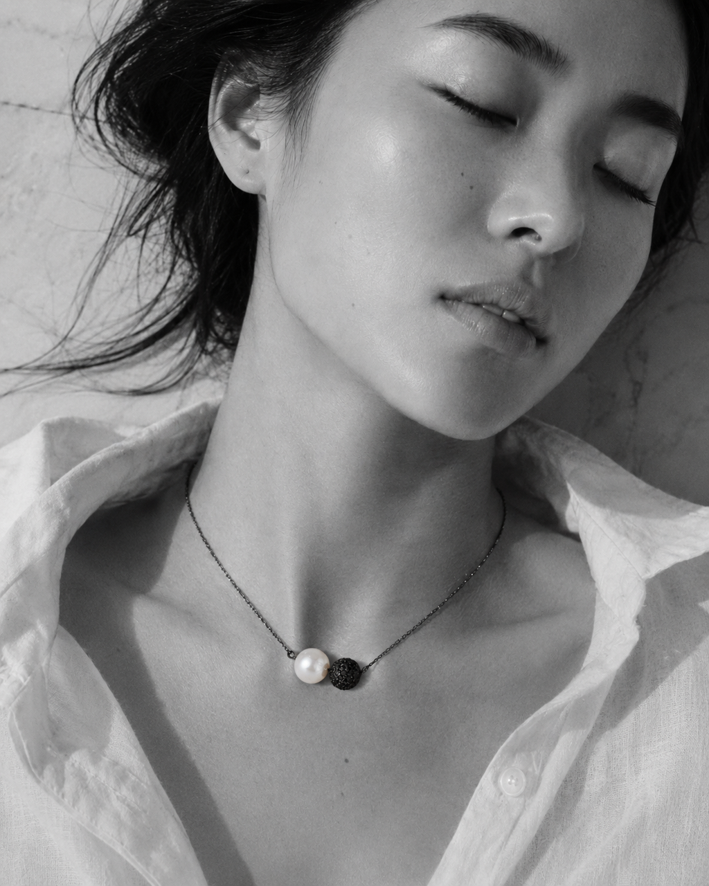 Zephyr pearl necklace black spinel oxidized silver worn by Asian model lying on marble, eyes closed, white linen shirt open, Mediterranean light, handmade by JLL Pearl
